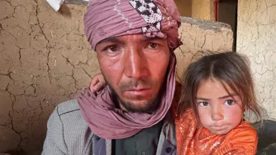Un hombre que lleva un turbante rosa abraza estrechamente a su pequeña hija frente a una pared de barro agrietada.